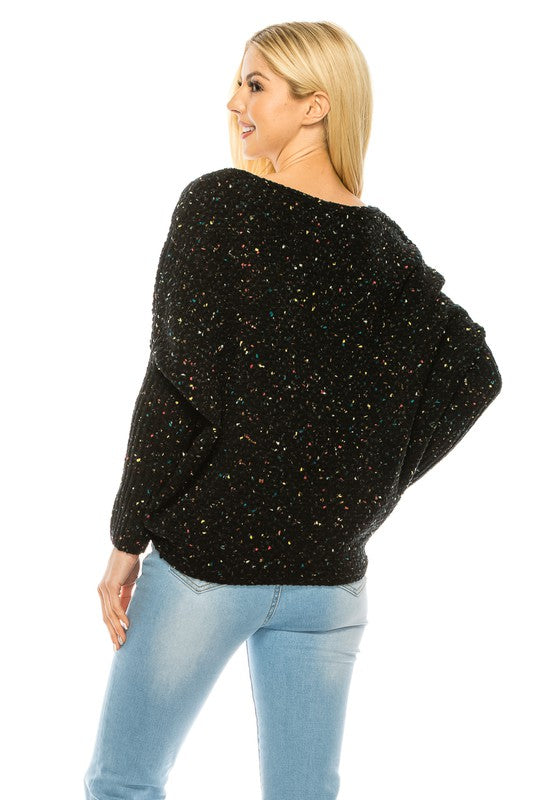 A woman with long blonde hair wears a multi-color speckled knit jumper and light blue jeans. She poses with one hand on her hip and a relaxed expression on her face.