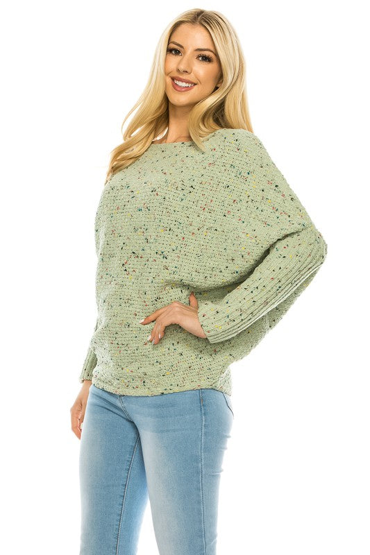 A woman with long blonde hair wears a multi-color speckled knit jumper and light blue jeans. She poses with one hand on her hip and a relaxed expression on her face.