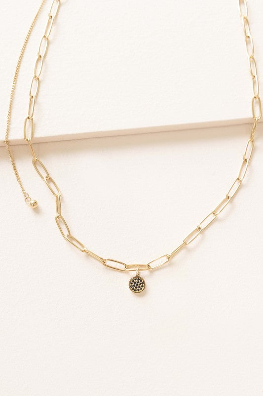 Cassio Charm Necklace featuring a gold double-chain design with a small black charm, embellished with black pavé stones, showcased on a light surface.