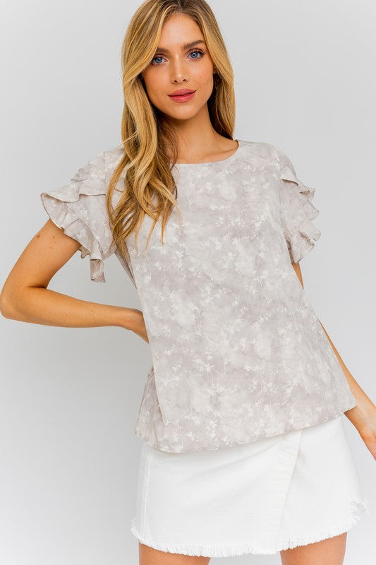 A woman in the S/S Overlap Ruffle Sleeve Ditsy Print Top and a white skirt stands against a plain background, exuding effortless elegance.