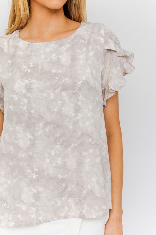 A woman in the S/S Overlap Ruffle Sleeve Ditsy Print Top and a white skirt stands against a plain background, exuding effortless elegance.