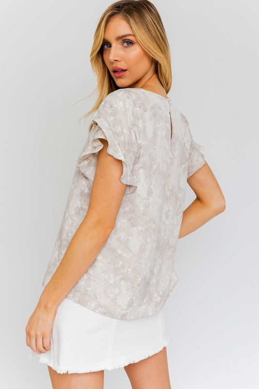 A woman in the S/S Overlap Ruffle Sleeve Ditsy Print Top and a white skirt stands against a plain background, exuding effortless elegance.