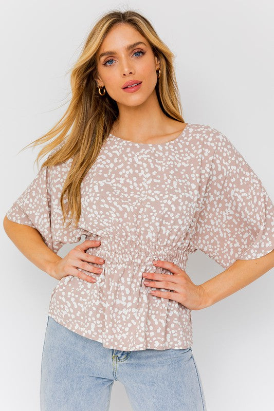 A woman poses with her hands on her hips against a plain background, wearing an abstract patterned top called the "Short Dolman Sleeve Smocking Waist Abstract Top" and blue jeans.