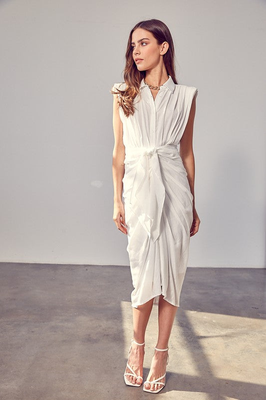 A woman in a white Sleeveless Button Front Tie Dress stands in a minimalist room with concrete flooring and neutral-colored walls. She has long brown hair and wears a necklace, making it perfect for summer gatherings.