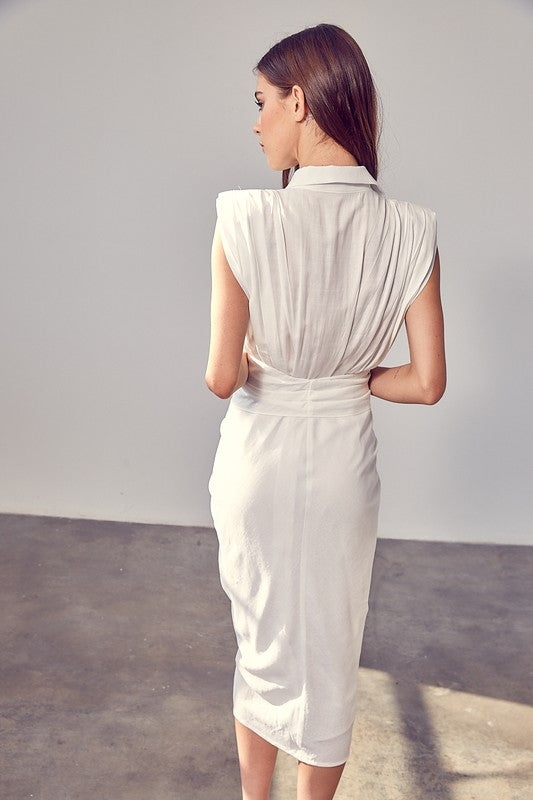 A woman in a white Sleeveless Button Front Tie Dress stands in a minimalist room with concrete flooring and neutral-colored walls. She has long brown hair and wears a necklace, making it perfect for summer gatherings.