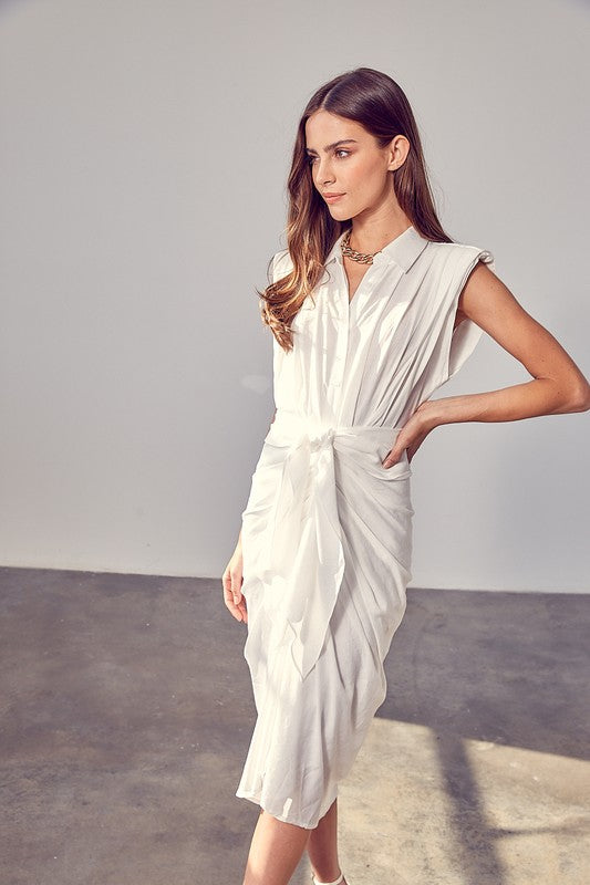 A woman in a white Sleeveless Button Front Tie Dress stands in a minimalist room with concrete flooring and neutral-colored walls. She has long brown hair and wears a necklace, making it perfect for summer gatherings.