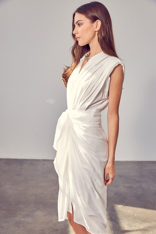 A woman in a white Sleeveless Button Front Tie Dress stands in a minimalist room with concrete flooring and neutral-colored walls. She has long brown hair and wears a necklace, making it perfect for summer gatherings.