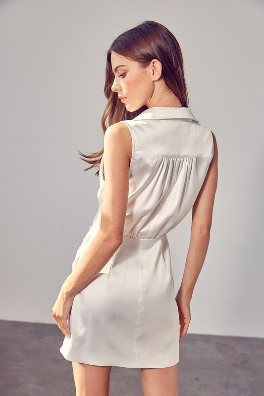 A woman in a sleeveless white Collar Wrap Dress stands in a bright room, looking to the side. She is wearing a gold chain necklace.