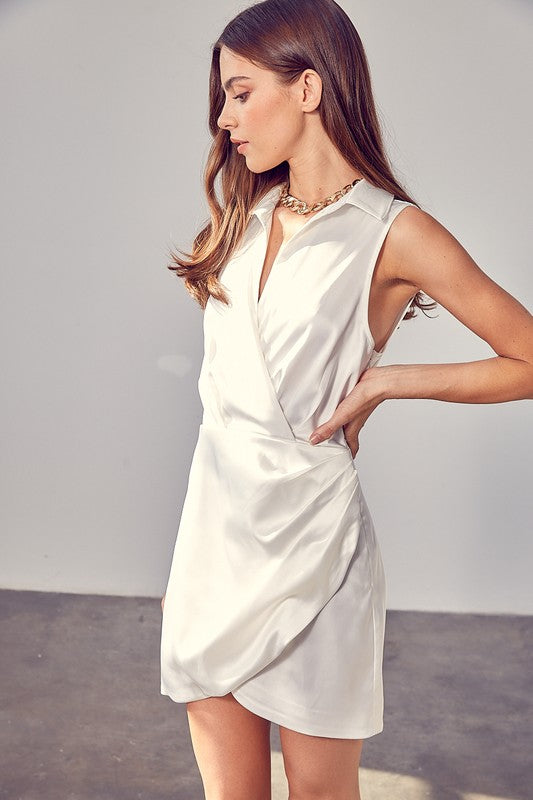 A woman in a sleeveless white Collar Wrap Dress stands in a bright room, looking to the side. She is wearing a gold chain necklace.