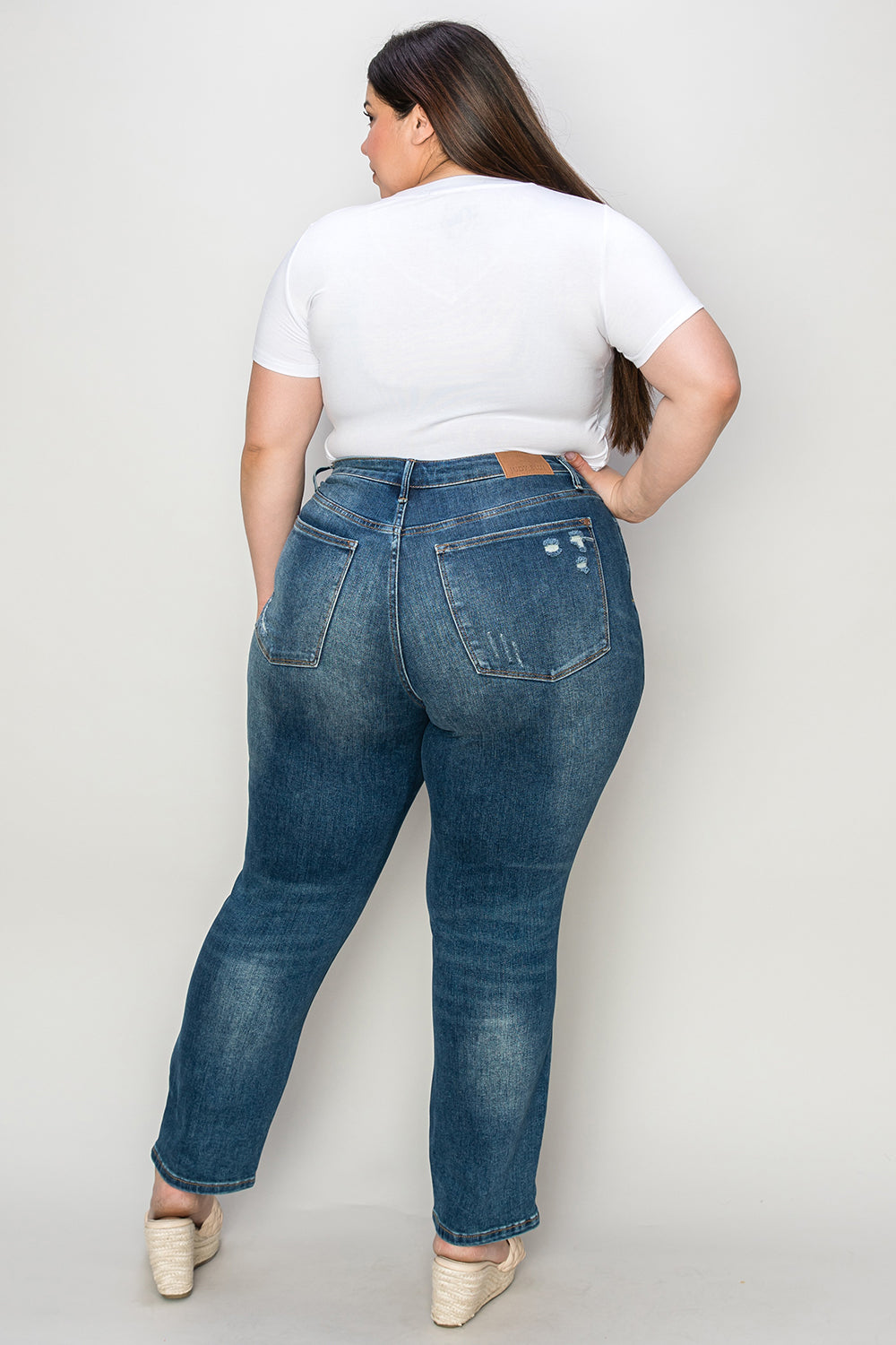 Person wearing Judy Blue Full Size Tummy Control High Waist Slim Jeans in blue and a white shirt, with their hands in their pockets. They are standing indoors on a polished concrete floor with a wooden cabinet and plant in the background.