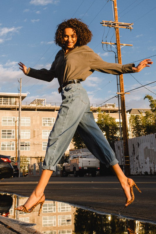 A woman wearing a gray shirt and Slouchy High Waisted Jeans, highlighted by front distress detailing, stands on a sunny street with her hands in her pockets. The background reveals an apartment building and utility poles, creating an atmosphere of laid-back comfort.