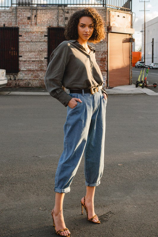 A woman wearing a gray shirt and Slouchy High Waisted Jeans, highlighted by front distress detailing, stands on a sunny street with her hands in her pockets. The background reveals an apartment building and utility poles, creating an atmosphere of laid-back comfort.