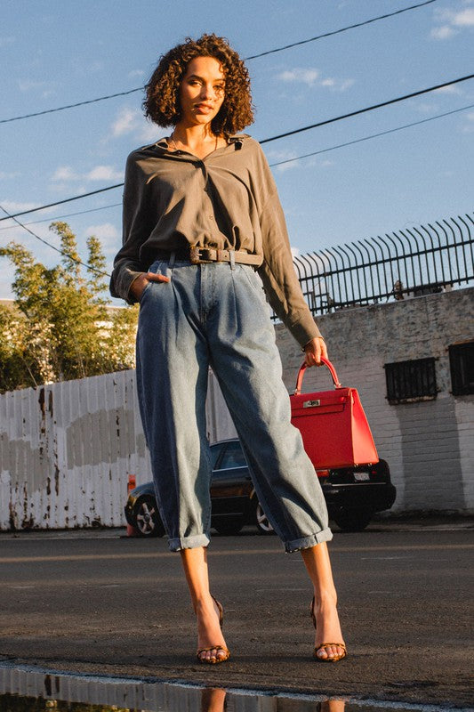 A woman wearing a gray shirt and Slouchy High Waisted Jeans, highlighted by front distress detailing, stands on a sunny street with her hands in her pockets. The background reveals an apartment building and utility poles, creating an atmosphere of laid-back comfort.