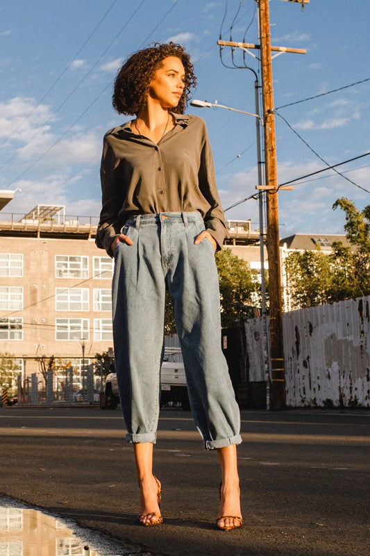 A woman wearing a gray shirt and Slouchy High Waisted Jeans, highlighted by front distress detailing, stands on a sunny street with her hands in her pockets. The background reveals an apartment building and utility poles, creating an atmosphere of laid-back comfort.