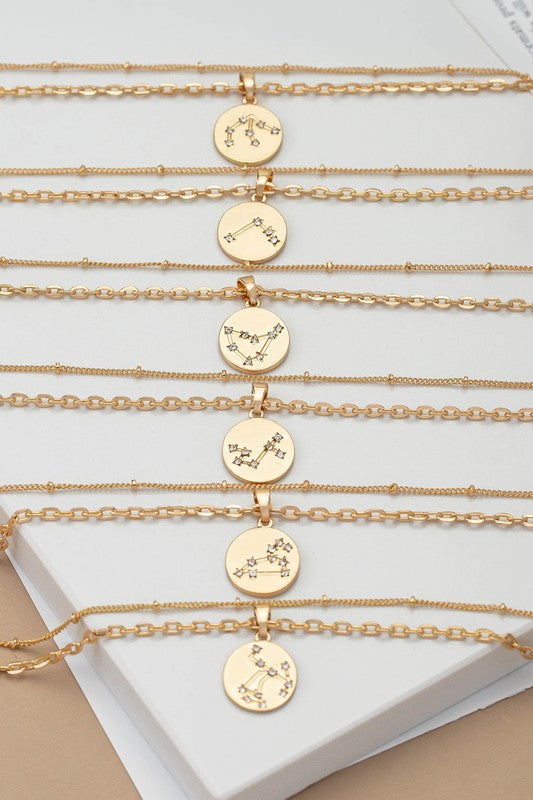 A display of gold-plated "zodiac sign pendant necklaces with rhinestones," featuring circular pendants adorned with different star constellations and zodiac sign motifs, arranged in rows on a white and beige surface.