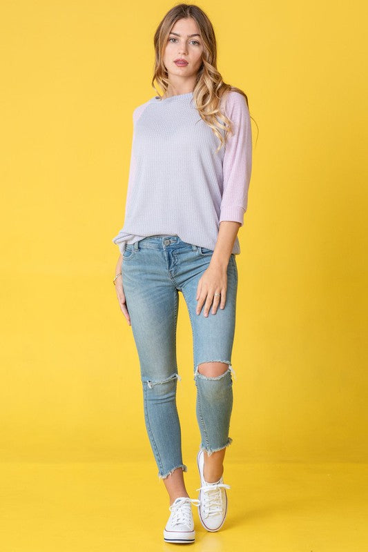 A person in a light blue Waffle Knit Two Tone Tunic and jeans stands in front of a yellow background, wearing a long necklace.