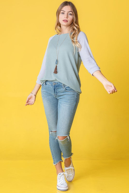 A woman with long hair wearing a Waffle Knit Two Tone Tunic in light blue and pink, paired with white pants, stands against a yellow background.