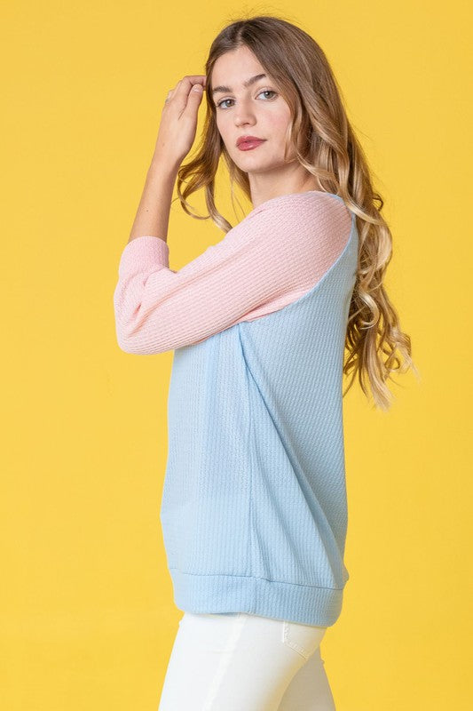 A woman with long hair wearing a Waffle Knit Two Tone Tunic in light blue and pink, paired with white pants, stands against a yellow background.