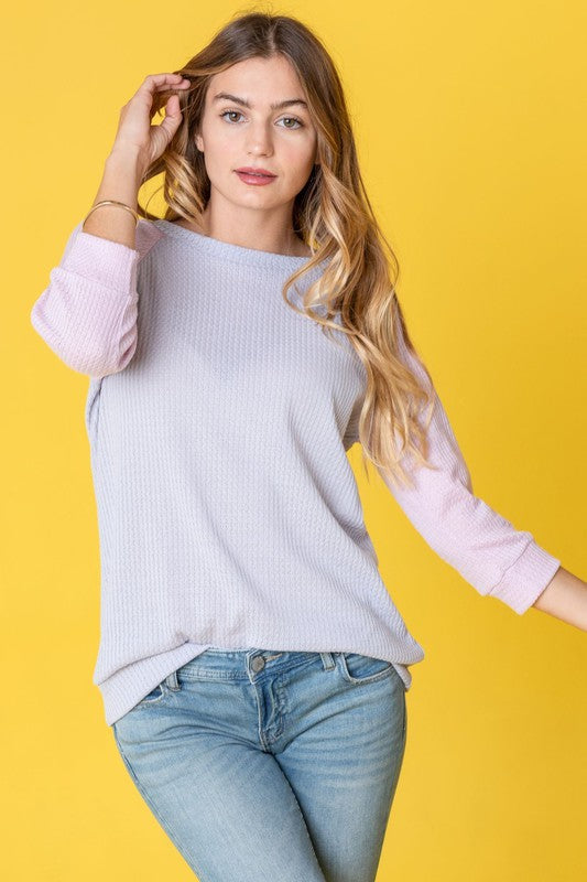 A woman with long hair wearing a Waffle Knit Two Tone Tunic in light blue and pink, paired with white pants, stands against a yellow background.