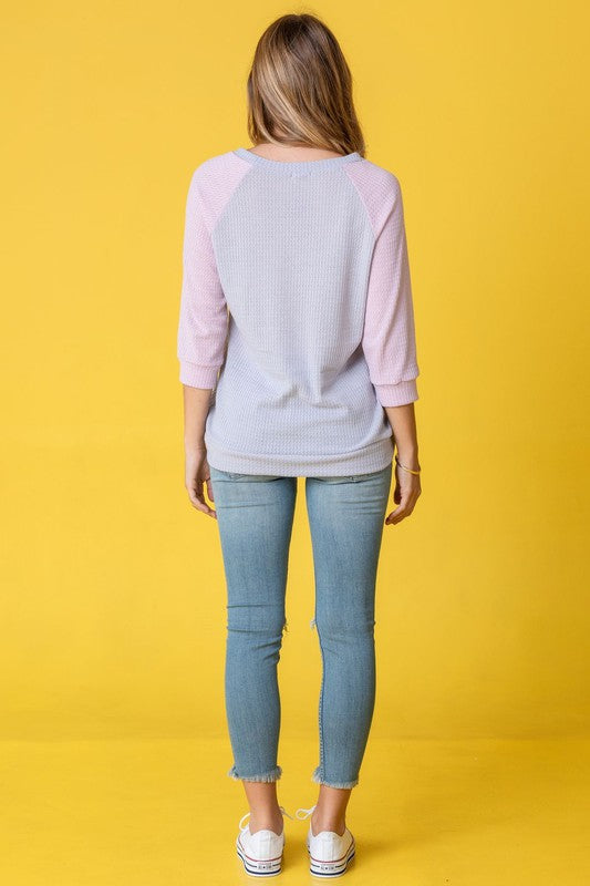 A person in a light blue Waffle Knit Two Tone Tunic and jeans stands in front of a yellow background, wearing a long necklace.