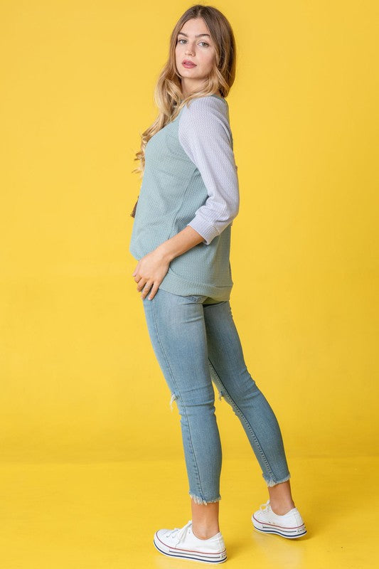 A woman with long hair wearing a Waffle Knit Two Tone Tunic in light blue and pink, paired with white pants, stands against a yellow background.