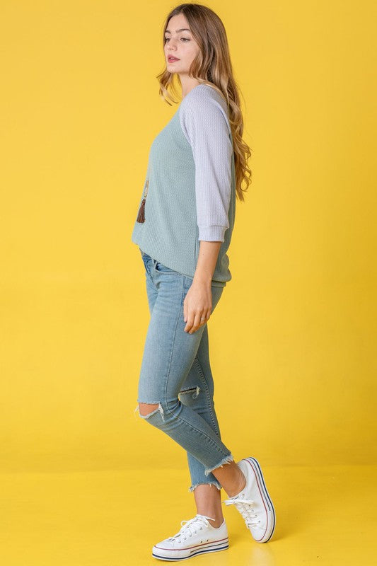 A person in a light blue Waffle Knit Two Tone Tunic and jeans stands in front of a yellow background, wearing a long necklace.