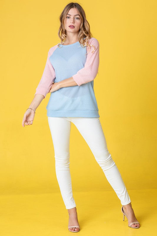 A woman with long hair wearing a Waffle Knit Two Tone Tunic in light blue and pink, paired with white pants, stands against a yellow background.