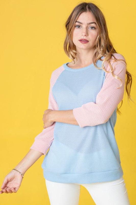 A woman with long hair wearing a Waffle Knit Two Tone Tunic in light blue and pink, paired with white pants, stands against a yellow background.