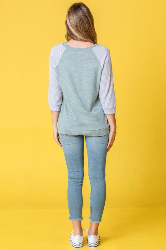 A person in a light blue Waffle Knit Two Tone Tunic and jeans stands in front of a yellow background, wearing a long necklace.