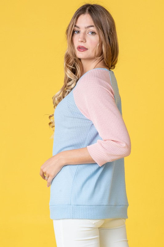 A woman with long hair wearing a Waffle Knit Two Tone Tunic in light blue and pink, paired with white pants, stands against a yellow background.