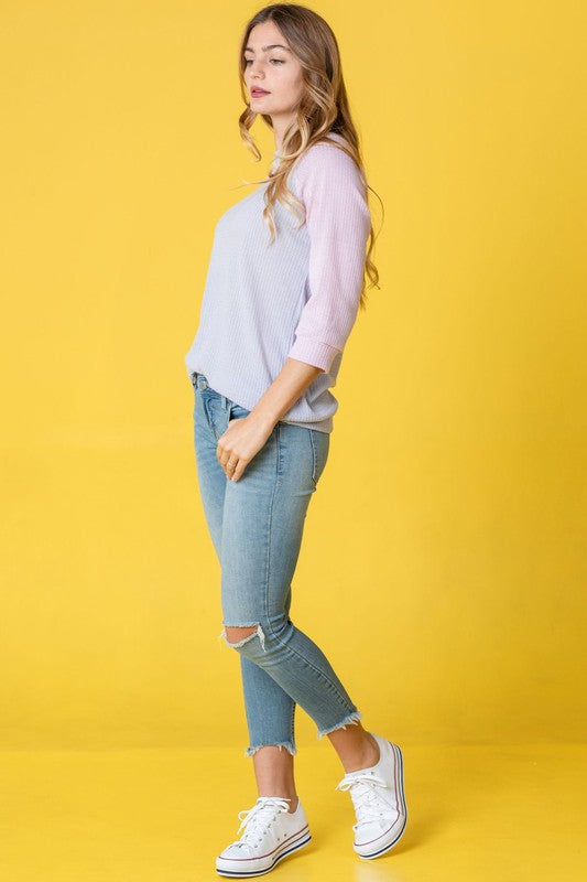 A person in a light blue Waffle Knit Two Tone Tunic and jeans stands in front of a yellow background, wearing a long necklace.