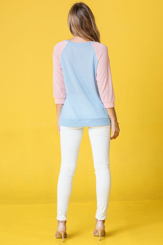 A woman with long hair wearing a Waffle Knit Two Tone Tunic in light blue and pink, paired with white pants, stands against a yellow background.