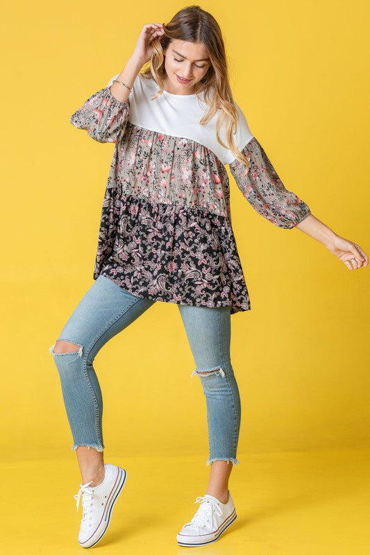 A person wearing a Three Tiered Bishop Sleeve Paisley Floral Tunic and jeans stands gracefully against a vivid yellow background.