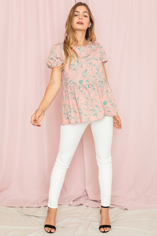 A woman wearing a Paisley Short Puff Sleeve Tunic and white pants stands against a pink backdrop, looking to the side.