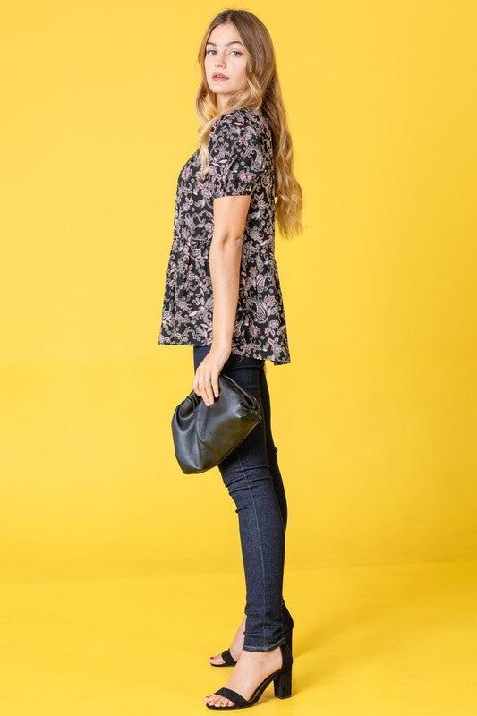 A woman wearing a Paisley Short Puff Sleeve Tunic and white pants stands against a pink backdrop, looking to the side.