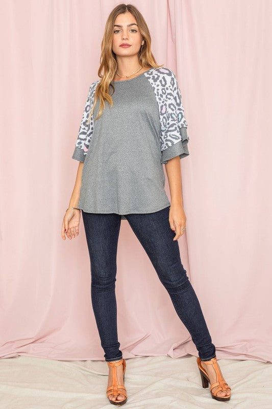 A woman wearing a Double Ruffle Sleeve Leopard Tunic poses gracefully in front of a pink curtain, showcasing her unique style.