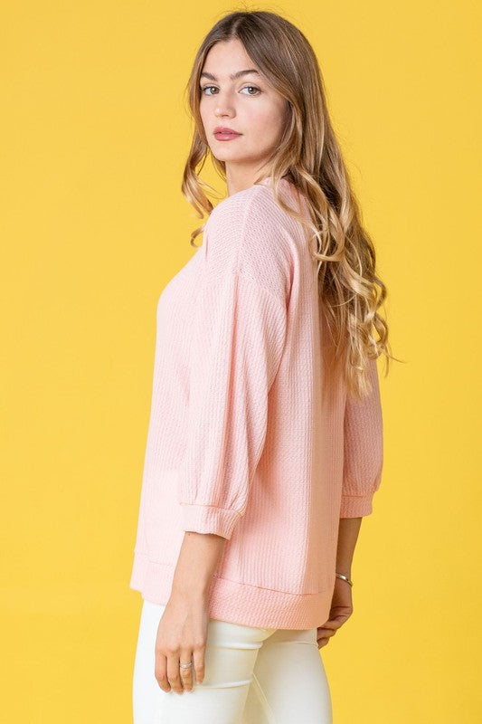 A person with long hair wearing a Solid Waffle Knit Top in pink, paired with white pants, stands against a yellow background.