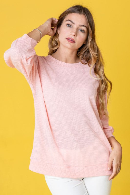 A person with long hair wearing a Solid Waffle Knit Top in pink, paired with white pants, stands against a yellow background.