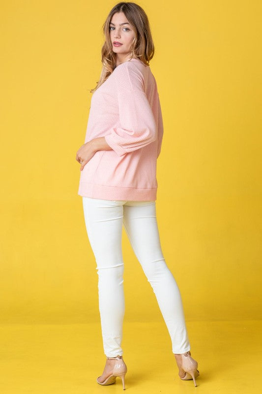 A person with long hair wearing a Solid Waffle Knit Top in pink, paired with white pants, stands against a yellow background.