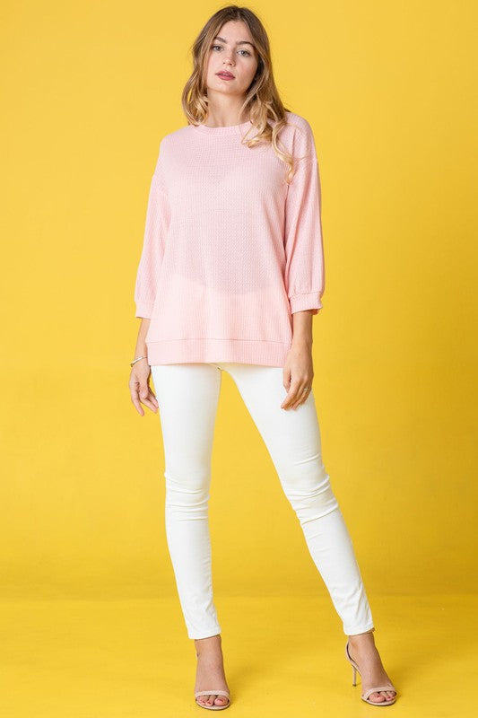 A person with long hair wearing a Solid Waffle Knit Top in pink, paired with white pants, stands against a yellow background.