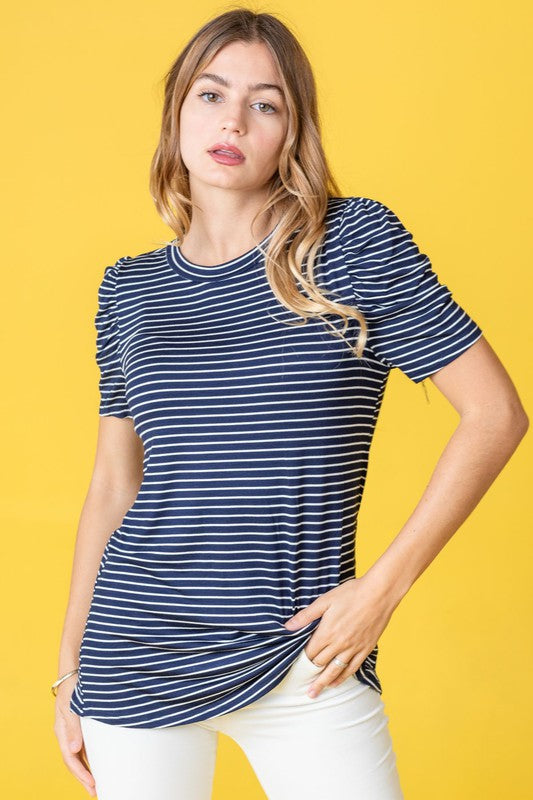 A woman in a Plus Stripe Puff Ruched Sleeve Top, featuring gray and white stripes, stands against a light pink backdrop. The top is made in the United States and has short ruched sleeves.