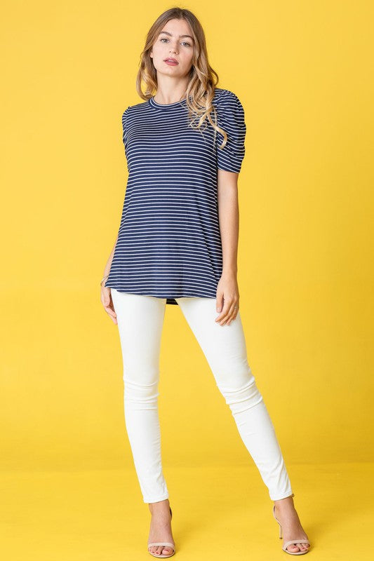 A woman presents the Stripe Puff Ruched Sleeve Top, a navy and white shirt with ruched sleeves, against a vibrant yellow background.