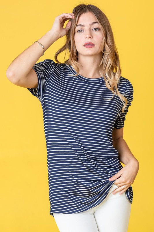 A woman in a Plus Stripe Puff Ruched Sleeve Top, featuring gray and white stripes, stands against a light pink backdrop. The top is made in the United States and has short ruched sleeves.