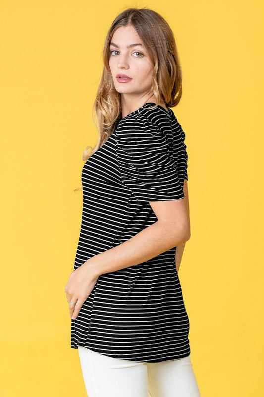 A woman in a Plus Stripe Puff Ruched Sleeve Top, featuring gray and white stripes, stands against a light pink backdrop. The top is made in the United States and has short ruched sleeves.