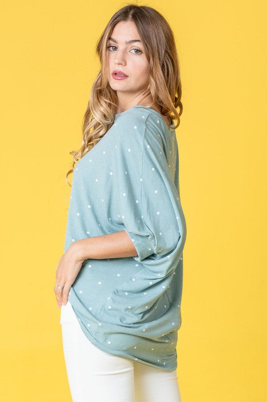 A person dressed in a pink Polka Dot Dolman Tunic and blue jeans stands in a room with wooden flooring and white walls.