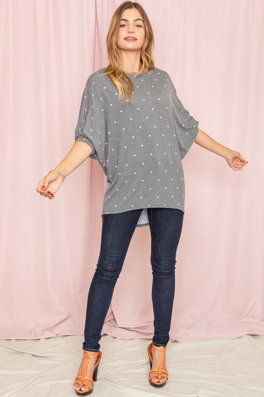 A person dressed in a pink Polka Dot Dolman Tunic and blue jeans stands in a room with wooden flooring and white walls.