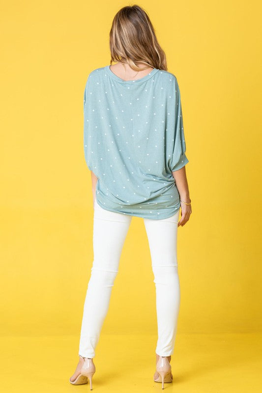 A person dressed in a pink Polka Dot Dolman Tunic and blue jeans stands in a room with wooden flooring and white walls.