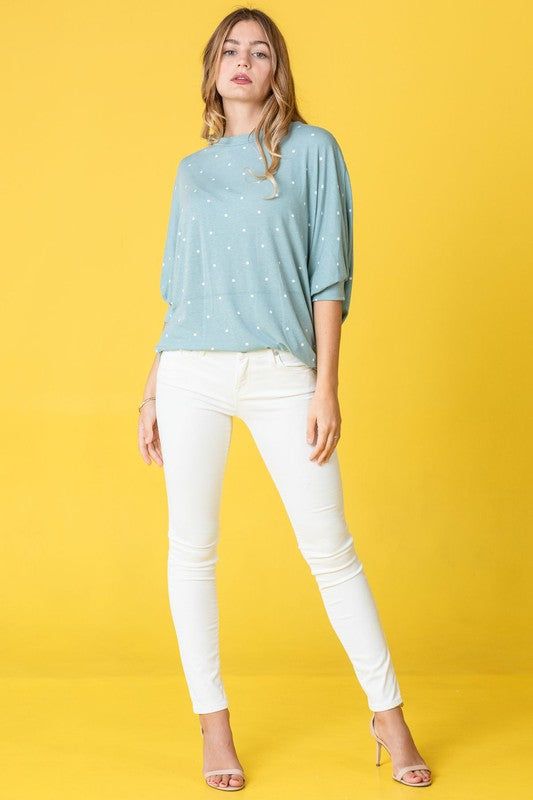 A person dressed in a pink Polka Dot Dolman Tunic and blue jeans stands in a room with wooden flooring and white walls.