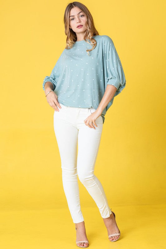 A person dressed in a pink Polka Dot Dolman Tunic and blue jeans stands in a room with wooden flooring and white walls.