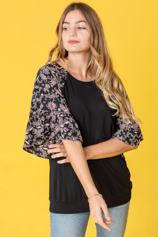 A woman stands against a vibrant yellow background wearing the Ruffle Print Sleeve Tunic, a chic black outfit combining elegance and playful charm. Made in the United States, it features stylish ruffle print sleeves.